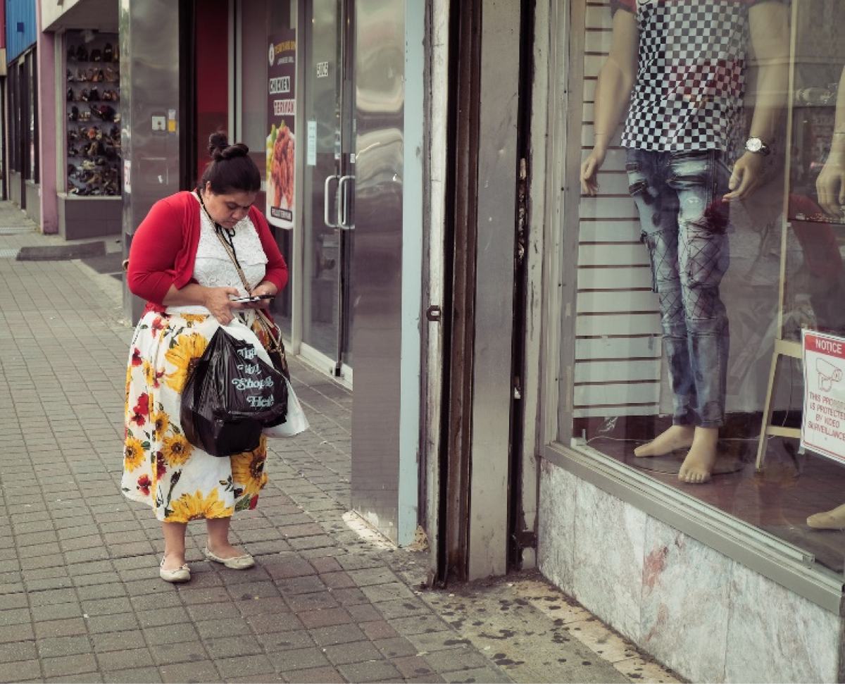 street photography, NY street photography, NY photography, americana photography, urban street life, woman, texting, on phone, near store, overweight, carrying bags, flower skirt, red coat, white shirt